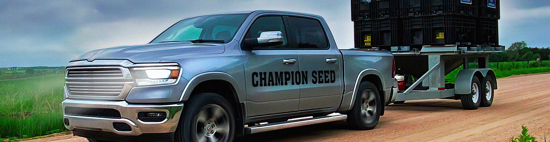 Truck with Champion Seed printed on the site, hauling a trailer that has two Champion Seed crates full of seed on top. The truck is driving down a dirt road with green grass and cloudly sky.